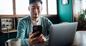 Confident young Asian man looking at smartphone while working on laptop computer in home office. Remote working, freelancer, small business concept